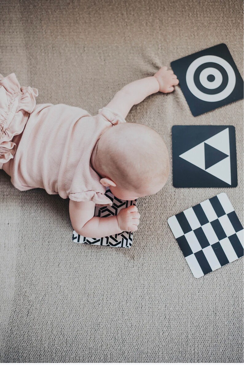 May include: A baby lying on its tummy on a beige carpet, reaching for a black and white card with a circle target design. Three other black and white cards are visible on the carpet, featuring a triangle, a checkerboard pattern, and a geometric design.