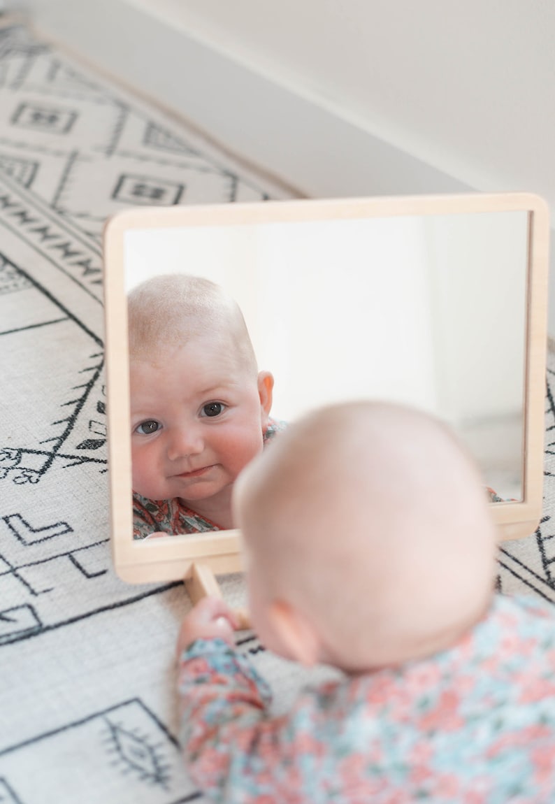 May include: A baby looking at their reflection in a wooden framed mirror. The baby is wearing a floral print onesie.