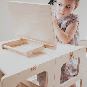 May include: A wooden step stool with a built-in easel and mirror. The easel has a wooden handle and the words "Jenny & Co." are printed on the side. A young child is sitting on the stool and looking in the mirror.