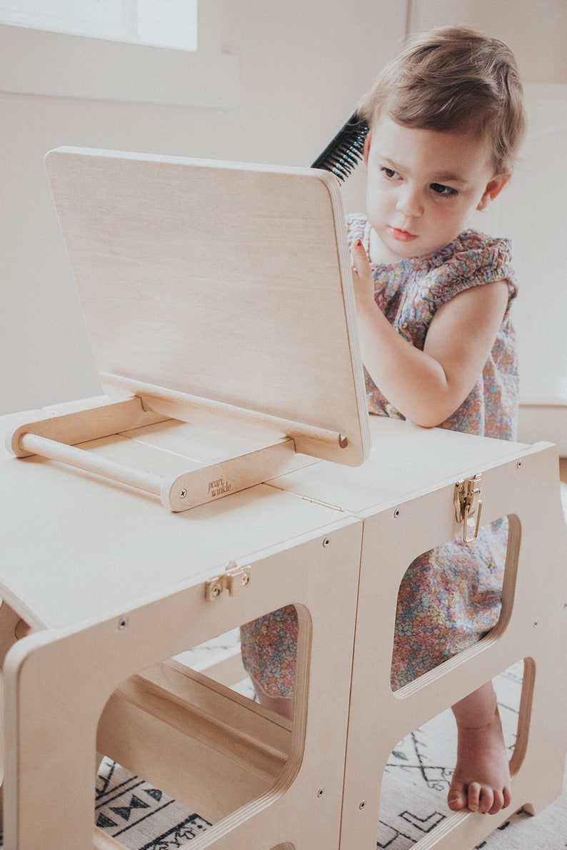 May include: A wooden step stool with a built-in easel and mirror. The easel has a wooden handle and the words "Jenny & Co." are printed on the side. A young child is sitting on the stool and looking in the mirror.