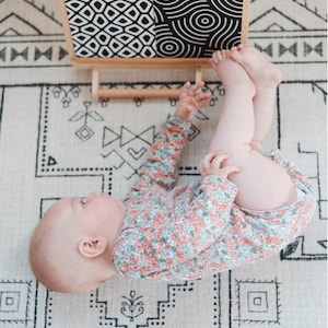 May include: A baby lying on a white and black patterned rug, looking up at a wooden activity board with black and white geometric patterns.