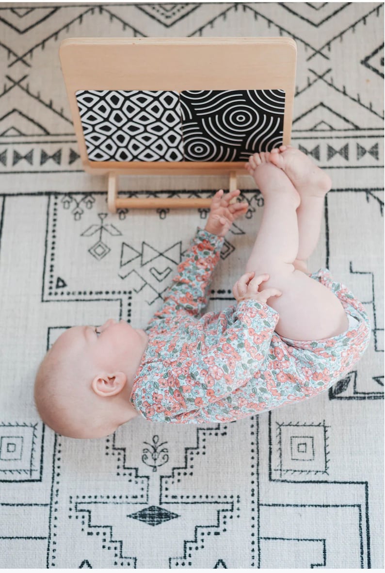 May include: A baby lying on a white and black patterned rug, looking up at a wooden activity board with black and white geometric patterns.