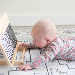 May include: A baby wearing a floral onesie is looking at a black and white patterned board with wooden handles. The board is propped up on a white and black patterned rug.