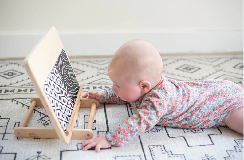 May include: A baby wearing a floral onesie is looking at a black and white patterned board with wooden handles. The board is propped up on a white and black patterned rug.