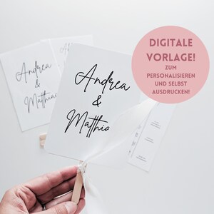 May include: A white wedding fan with the names "Andrea & Matthias" written in black calligraphy. The fan is on a wooden stick and has a white ribbon tied around it. The text "DIGITALE VORLAGE! ZUM PERSONALISIEREN UND SELBST AUSDRUCKEN!" is written in pink on a white circle.