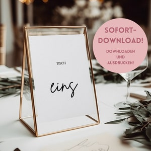May include: A gold metal table number holder with a white card that says "Tisch eins" in black cursive writing. The card is in a gold frame. The background is a blurry image of a table setting with greenery.