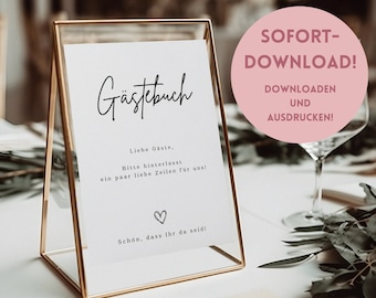 Puede incluir: Un cartel con marco dorado, fondo blanco y texto negro que dice "Gästebuch" (alemán para "Libro de visitas"). El texto debajo dice "Liebe Gäste, Bitte hinterlasst ein paar liebe Zeilen für uns!" (alemán para "Estimados invitados, por favor, dejen algunas palabras amables para nosotros!") y "Schön, dass Ihr da seid!" (alemán para "¡Es agradable que estéis aquí!"). El cartel está sobre una mesa con un vaso de agua y vegetación.