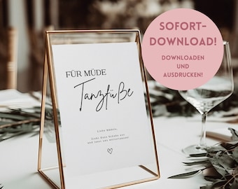 Puede incluir: Un cartel imprimible en un marco dorado que dice "FÜR MÜDE Tanzfüße" con el texto "Liebe Madels, Zieht Eure Schuhe aus und lasst uns weiter tanzen!" y un símbolo de corazón. El cartel está sobre una mesa con un vaso de agua y vegetación.
