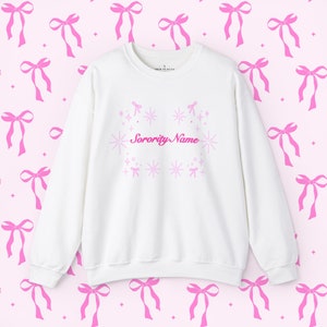 Könnte beinhalten: Weißes Sweatshirt mit der Aufschrift "Sorority Name" in rosa Schrift, umgeben von rosa Sternen und Schleifen. Der Hintergrund zeigt ein sich wiederholendes Muster aus rosa Schleifen auf einer hellrosa Oberfläche.