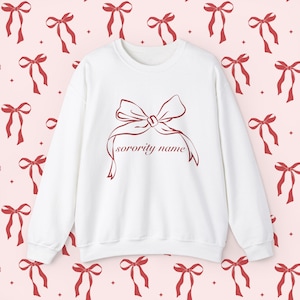 Puede incluir: Sudadera blanca de cuello redondo con un gran lazo granate y el texto "sorority name". El fondo presenta un patrón de lazos granates sobre un fondo rosa. Una parte superior informal y cómoda.