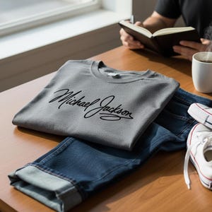 May include: A gray t-shirt with the name "Michael Jackson" in black script, folded on a wooden table next to blue jeans and white sneakers. A person is reading a book in the background.