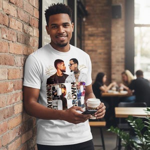 May include: White t-shirt featuring a graphic design with the text "Raymond & Brown" and images of two men. The shirt is being worn by a person. The person is holding a coffee cup and a phone.