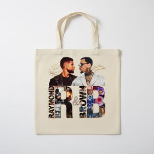 May include: A beige tote bag featuring a graphic design. The design includes images of two men, with the words "Raymond" and "Brown" on either side of the "&" symbol. The words "R&B" are also visible in the design.