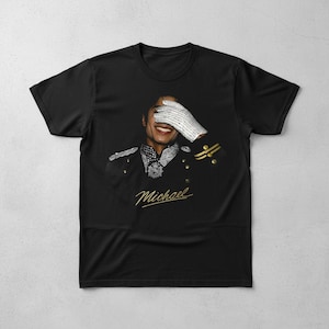 May include: Black t-shirt featuring a portrait of a smiling person wearing a military-style jacket and a white, bejeweled glove covering their eyes. The word "Michael" is written in gold script below the portrait.