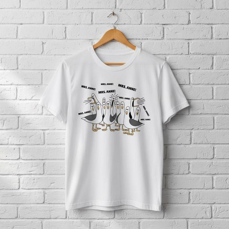 May include: White t-shirt featuring a cartoon illustration of seagulls with the text "MRS. ANNE!". The seagulls are in various poses, with black and white bodies, yellow feet, and open beaks. The shirt is hanging on a wooden hanger.