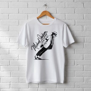 May include: White t-shirt featuring a black graphic of Michael Jackson in his signature lean pose, with his name in cursive script. The shirt is hanging on a wooden hanger against a white brick wall.