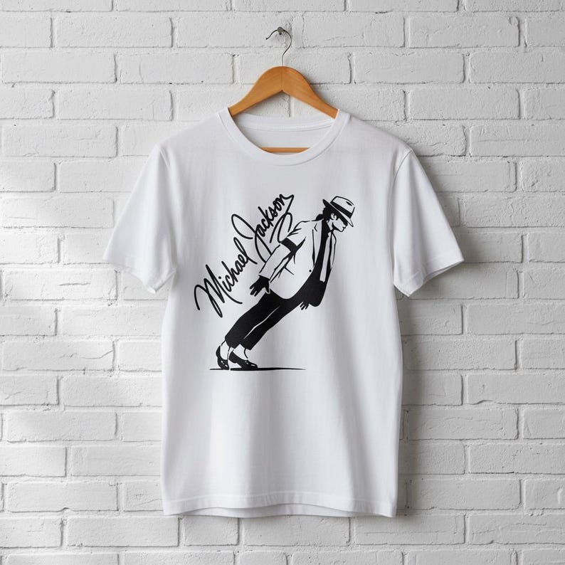 May include: White t-shirt featuring a black graphic of Michael Jackson in his signature lean pose, with his name in cursive script. The shirt is hanging on a wooden hanger against a white brick wall.