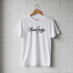 May include: White t-shirt with the black cursive text "Michael Jackson". The shirt is hanging on a wooden hanger against a white brick wall. The t-shirt is a classic crew neck style.