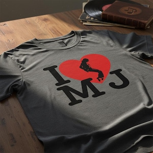 May include: Gray t-shirt with a red heart graphic featuring a silhouette of a person and the text "I MJ" in black. The shirt is displayed on a wooden surface, with vinyl records and a book in the background.