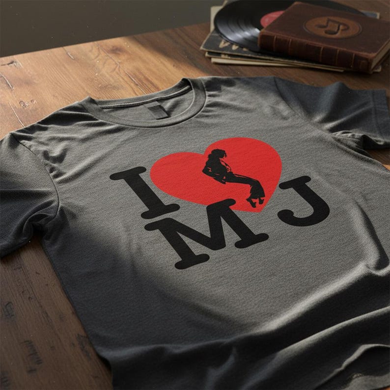May include: Gray t-shirt with a red heart graphic featuring a silhouette of a person and the text "I MJ" in black. The shirt is displayed on a wooden surface, with vinyl records and a book in the background.