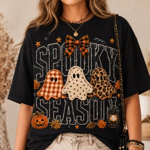 May include: A black short-sleeve t-shirt featuring the text "SPOOKY SEASON" in a distressed font. The design includes three ghosts with different patterns, an orange and black plaid bow, a jack-o'-lantern, autumn leaves, and a lantern.