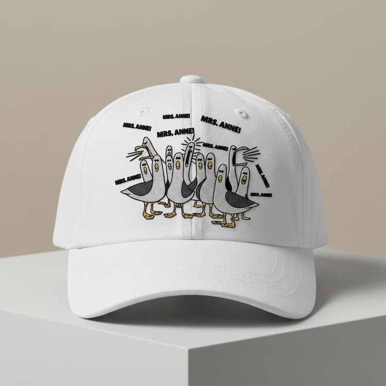 May include: White baseball cap featuring a cartoon illustration of seagulls with the text "MRS. ANNE!" in black. The seagulls are depicted in black and white with yellow feet and beaks. The cap has a curved brim and a button on top.