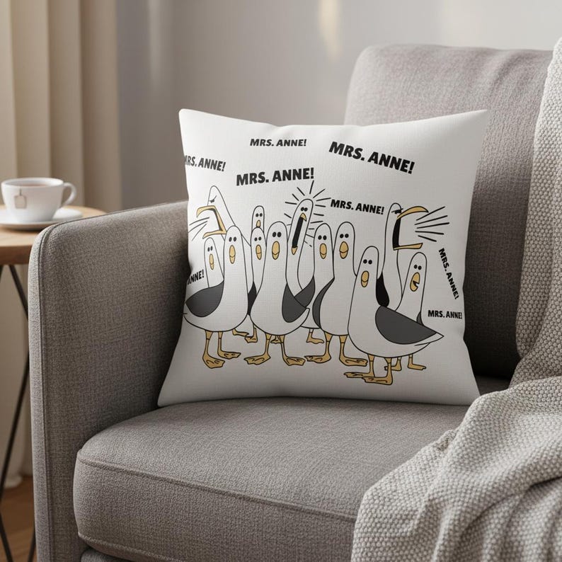 May include: White throw pillow featuring a cartoon illustration of seagulls with black and gray wings and yellow feet. The seagulls are yelling, with the text "MRS. ANNE!" repeated around them. The pillow is on a gray couch.