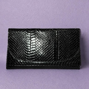 May include: Black clutch bag with a reptile skin effect. The rectangular bag has a flap closure and a subtle sheen. The accessory is set against a light purple background. The clutch is a fashion accessory.