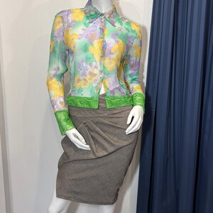 Op de afbeelding: Een mannequin met een zwart leren kattenmasker, een doorschijnende blouse met bloemenprint en groene kanten bies, en een bruine rok. De blouse heeft gele, paarse en groene bloempatronen. De rok is neutraal bruin van kleur.