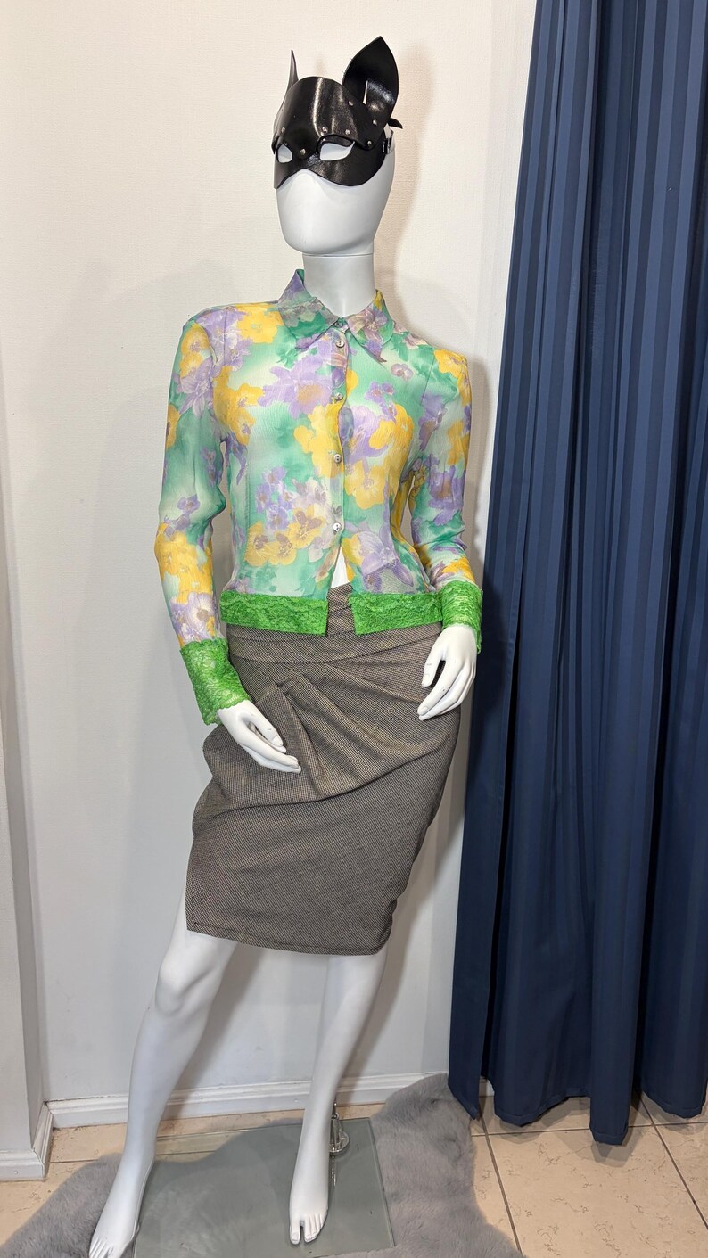 Op de afbeelding: Een mannequin met een zwart leren kattenmasker, een doorschijnende blouse met bloemenprint en groene kanten bies, en een bruine rok. De blouse heeft gele, paarse en groene bloempatronen. De rok is neutraal bruin van kleur.