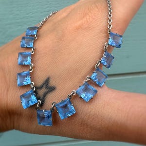 Op de afbeelding: Een zilverkleurige ketting met vierkante, lichtblauwe edelstenen. De ketting heeft een fijne ketting met de edelstenen gelijkmatig verdeeld. De edelstenen zijn helderblauw van kleur, wat een vleugje kleur aan het ontwerp geeft.