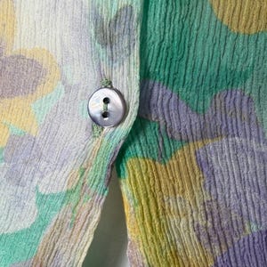 Op de afbeelding: Close-up van een bloemmotiefstof in pastelkleuren. De stof heeft een knoopsluiting met een parelmoeren knoop. Het ontwerp bevat tinten groen, geel en paars, waardoor een zacht, gestructureerd uiterlijk ontstaat. De onderrand onthult een zwart-wit pied-de-poule patroon.