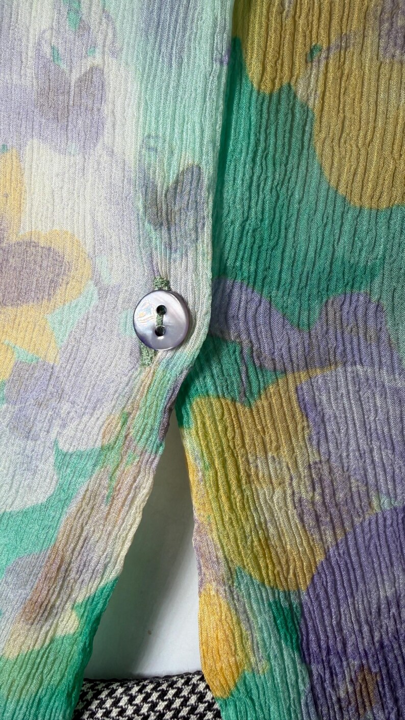 Op de afbeelding: Close-up van een bloemmotiefstof in pastelkleuren. De stof heeft een knoopsluiting met een parelmoeren knoop. Het ontwerp bevat tinten groen, geel en paars, waardoor een zacht, gestructureerd uiterlijk ontstaat. De onderrand onthult een zwart-wit pied-de-poule patroon.