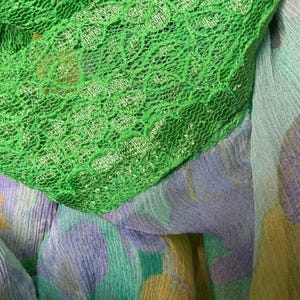 Op de afbeelding: Close-up van groene kanten stof gedrapeerd over een patroon sjaal. De sjaal heeft een aquarelontwerp met tinten groen, paars en geel. De texturen van de stoffen zijn zichtbaar, waardoor een gelaagd effect ontstaat.