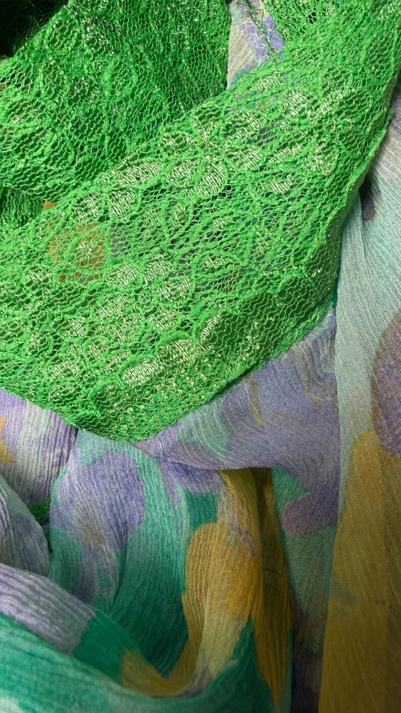 Op de afbeelding: Close-up van groene kanten stof gedrapeerd over een patroon sjaal. De sjaal heeft een aquarelontwerp met tinten groen, paars en geel. De texturen van de stoffen zijn zichtbaar, waardoor een gelaagd effect ontstaat.