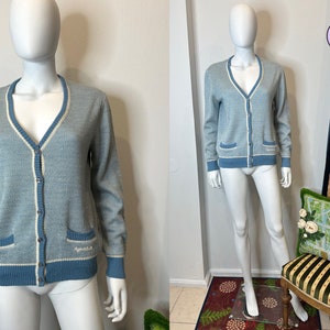 May include: A light blue cardigan with white trim and two pockets. The cardigan has a button closure and is made of a soft, knitted fabric.