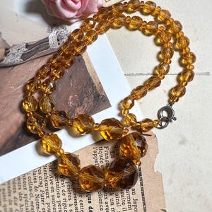 Puede incluir: Un collar vintage de cuentas de color ámbar con un cierre plateado. El collar presenta cuentas facetadas de varios tamaños, creando un efecto brillante. Una rosa rosa y un fondo de papel vintage también son visibles.
