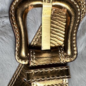 May include: Gold-toned leather belt with a prominent buckle featuring the letter "F". The belt has a metallic sheen and is stitched with a contrasting thread. The buckle is rectangular and has a gold finish.