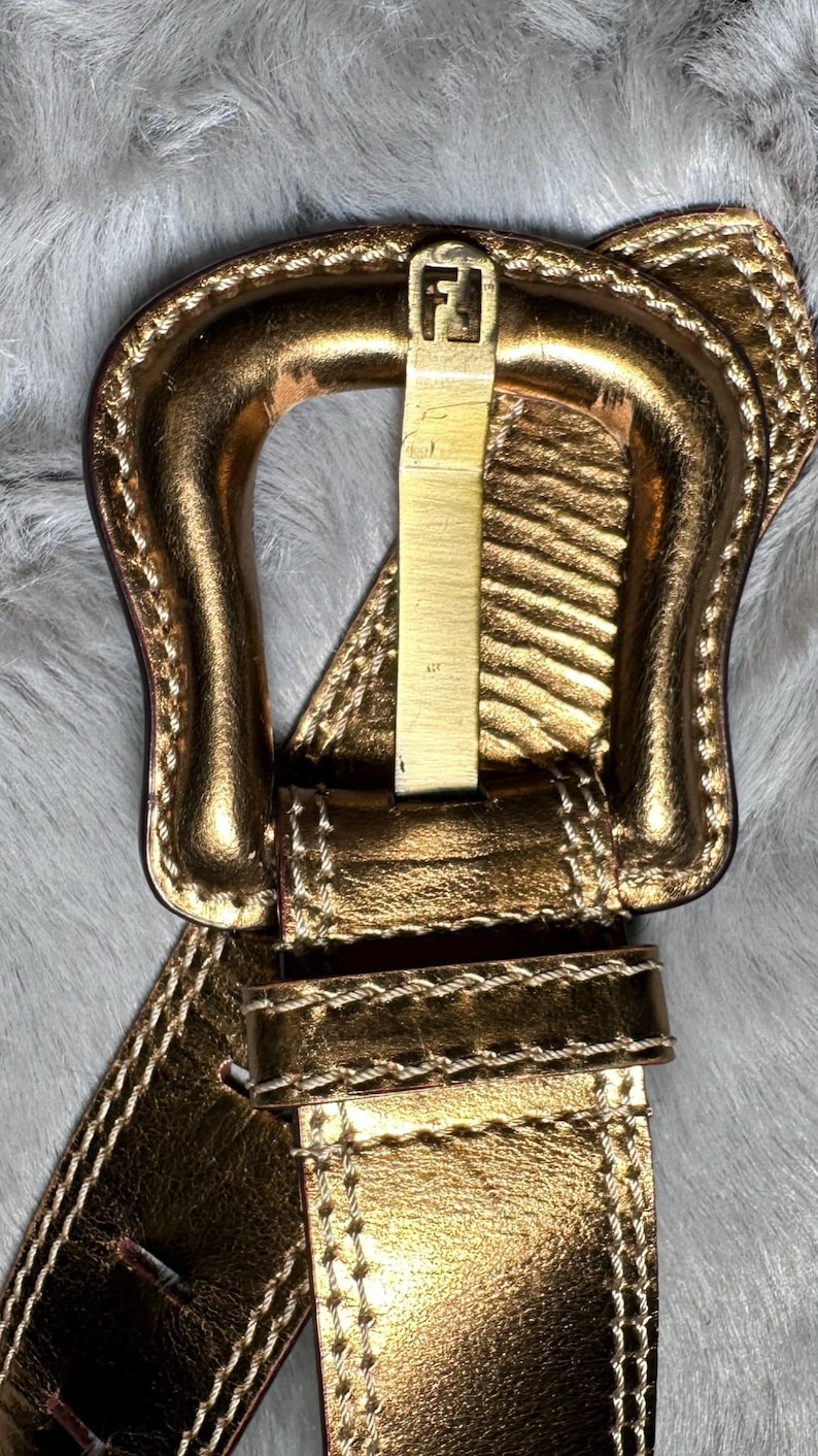 May include: Gold-toned leather belt with a prominent buckle featuring the letter "F". The belt has a metallic sheen and is stitched with a contrasting thread. The buckle is rectangular and has a gold finish.