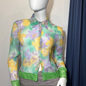 Op de afbeelding: Een doorschijnende blouse met knopen en een bloemenpatroon in geel, paars en groen. De blouse heeft lange mouwen met groene kanten manchetten en een groene kanten rand aan de taille. Een zwart leren kattenmasker wordt ook getoond.