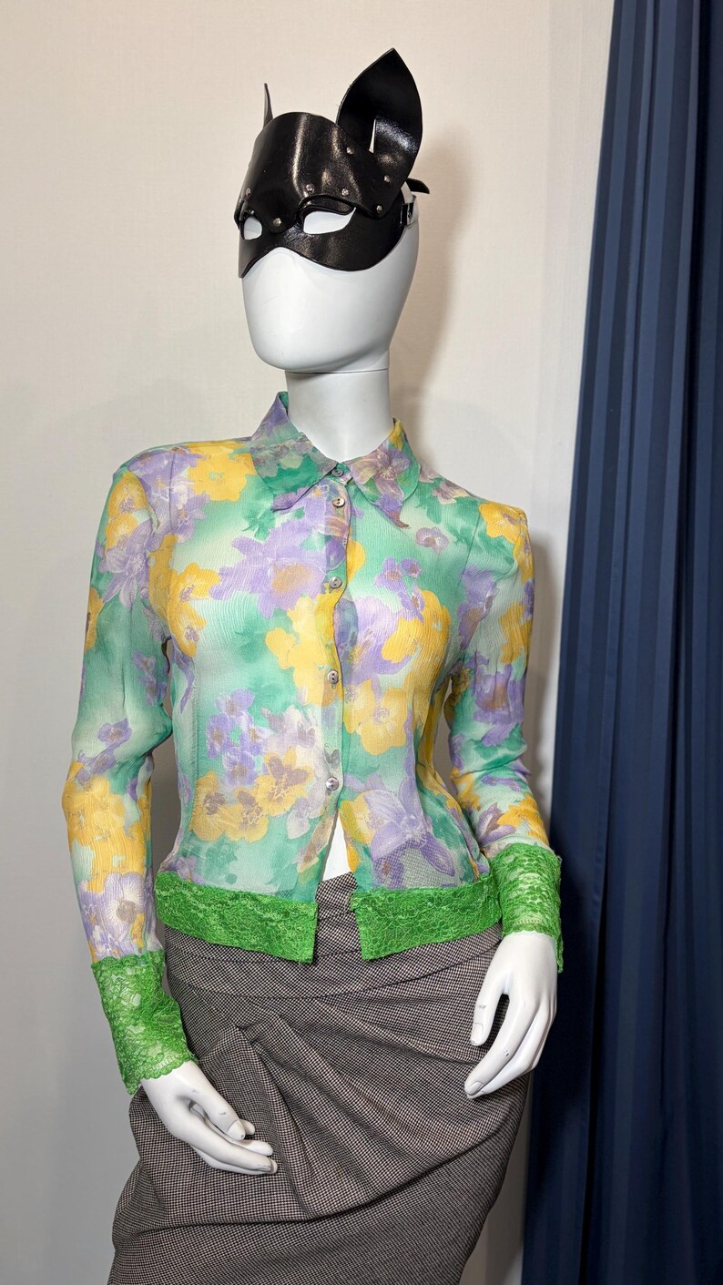 Op de afbeelding: Een doorschijnende blouse met knopen en een bloemenpatroon in geel, paars en groen. De blouse heeft lange mouwen met groene kanten manchetten en een groene kanten rand aan de taille. Een zwart leren kattenmasker wordt ook getoond.