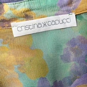 Op de afbeelding: Close-up van een bloemmotiefstof in groen, geel en paars. De stof heeft een gekreukte textuur en een label met de tekst "cristina capucci". Het kledingstuk lijkt een blouse of overhemd te zijn.