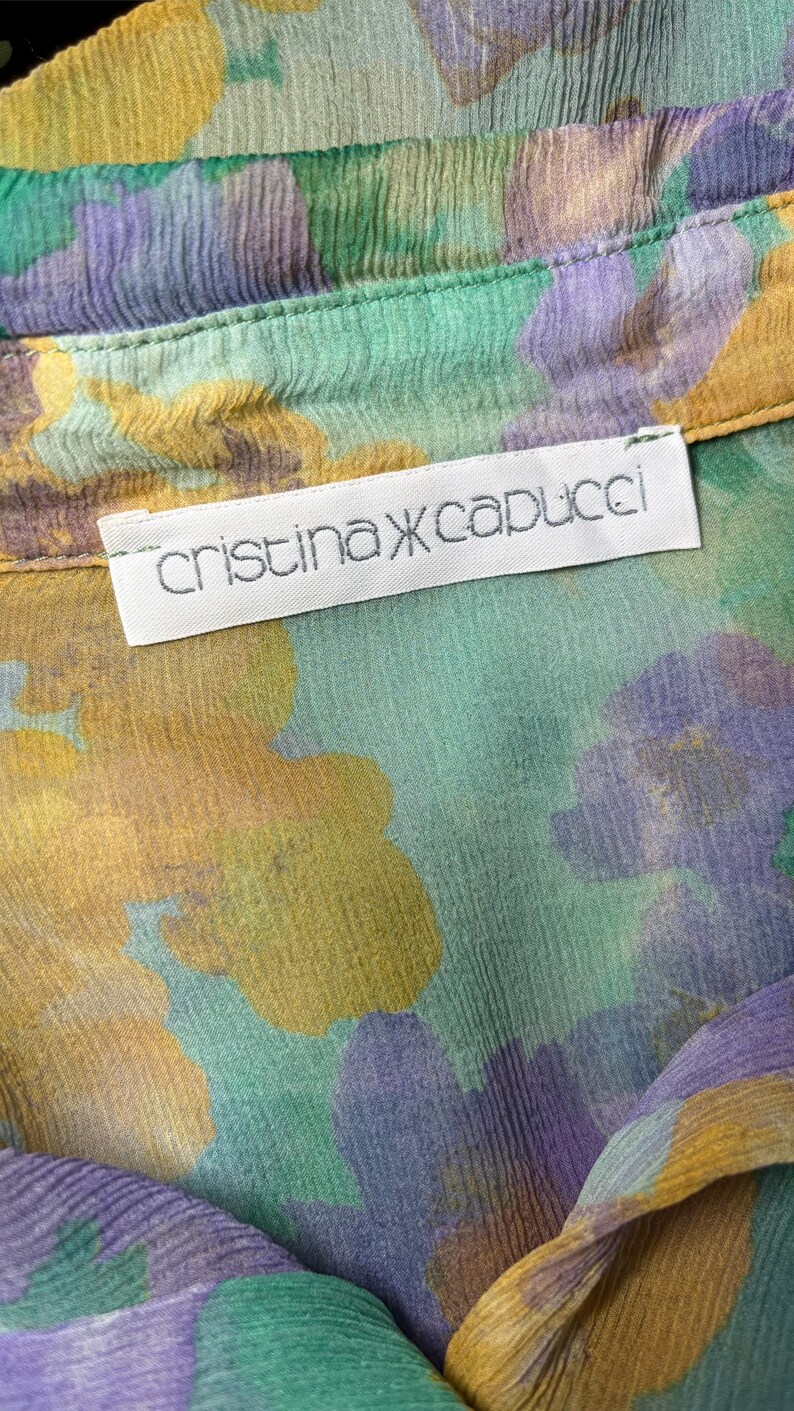 Op de afbeelding: Close-up van een bloemmotiefstof in groen, geel en paars. De stof heeft een gekreukte textuur en een label met de tekst "cristina capucci". Het kledingstuk lijkt een blouse of overhemd te zijn.