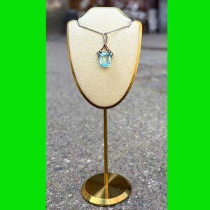 Op de afbeelding: Een zilverkleurige ketting met een rechthoekige, lichtblauwe gefacetteerde edelsteen hanger. De hanger hangt aan een dunne zilveren ketting en wordt getoond op een crèmekleurige, schildvormige standaard met een goudkleurige metalen voet.