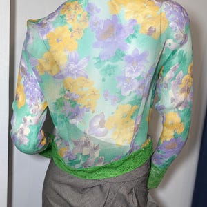 Op de afbeelding: Een doorschijnende blouse met lange mouwen met een bloemenpatroon in geel, paars en wit op een mintgroene achtergrond. De blouse wordt gecombineerd met een grijze rok en een groene bies aan de taille en manchetten. De mannequin draagt een rok.