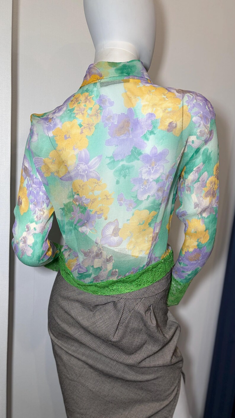 Op de afbeelding: Een doorschijnende blouse met lange mouwen met een bloemenpatroon in geel, paars en wit op een mintgroene achtergrond. De blouse wordt gecombineerd met een grijze rok en een groene bies aan de taille en manchetten. De mannequin draagt een rok.