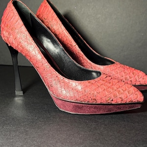 Puede incluir: Un par de zapatos de tacón alto rojos con un patrón texturizado. Los zapatos tienen tacones negros y una plataforma burdeos. El interior de los zapatos es negro.