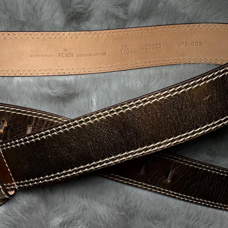 May include: Close-up of two brown leather belts with white stitching. One belt is stamped with "MADE IN ITALY", "FENDI", and "GENUINE LEATHER". The other belt is a darker brown with white stitching. The belts are on a gray, textured surface.