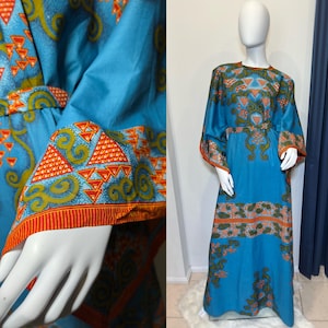 Op de afbeelding: Een lange, vloeiende blauwe jurk met een levendig patroon. De jurk heeft een ronde halslijn, lange mouwen en een getailleerde taille. De mouwen en zoom zijn afgezet met oranje en groene geometrische ontwerpen. De jurk wordt op een mannequin getoond.