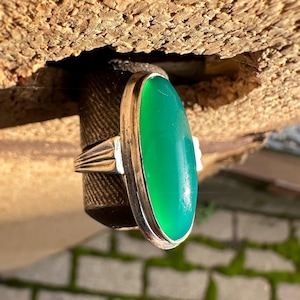 May include: A silver ring with a large, oval, bright green gemstone. The ring's band has a ribbed design on the sides. The gemstone is set in a silver bezel. The ring is displayed against a textured, brown background.